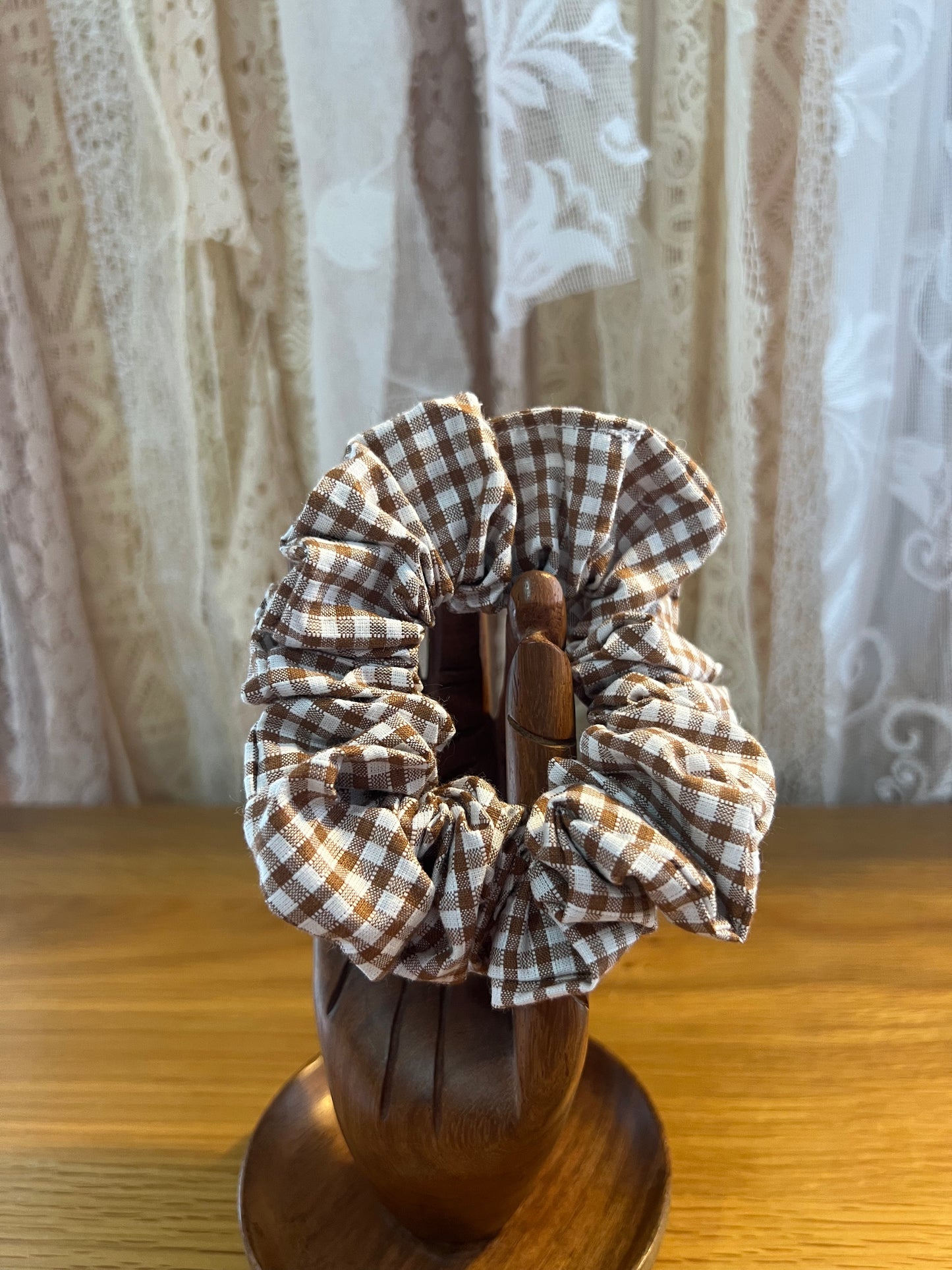 Brown checkered scrunchie on a wooden holder with a lace curtain background
