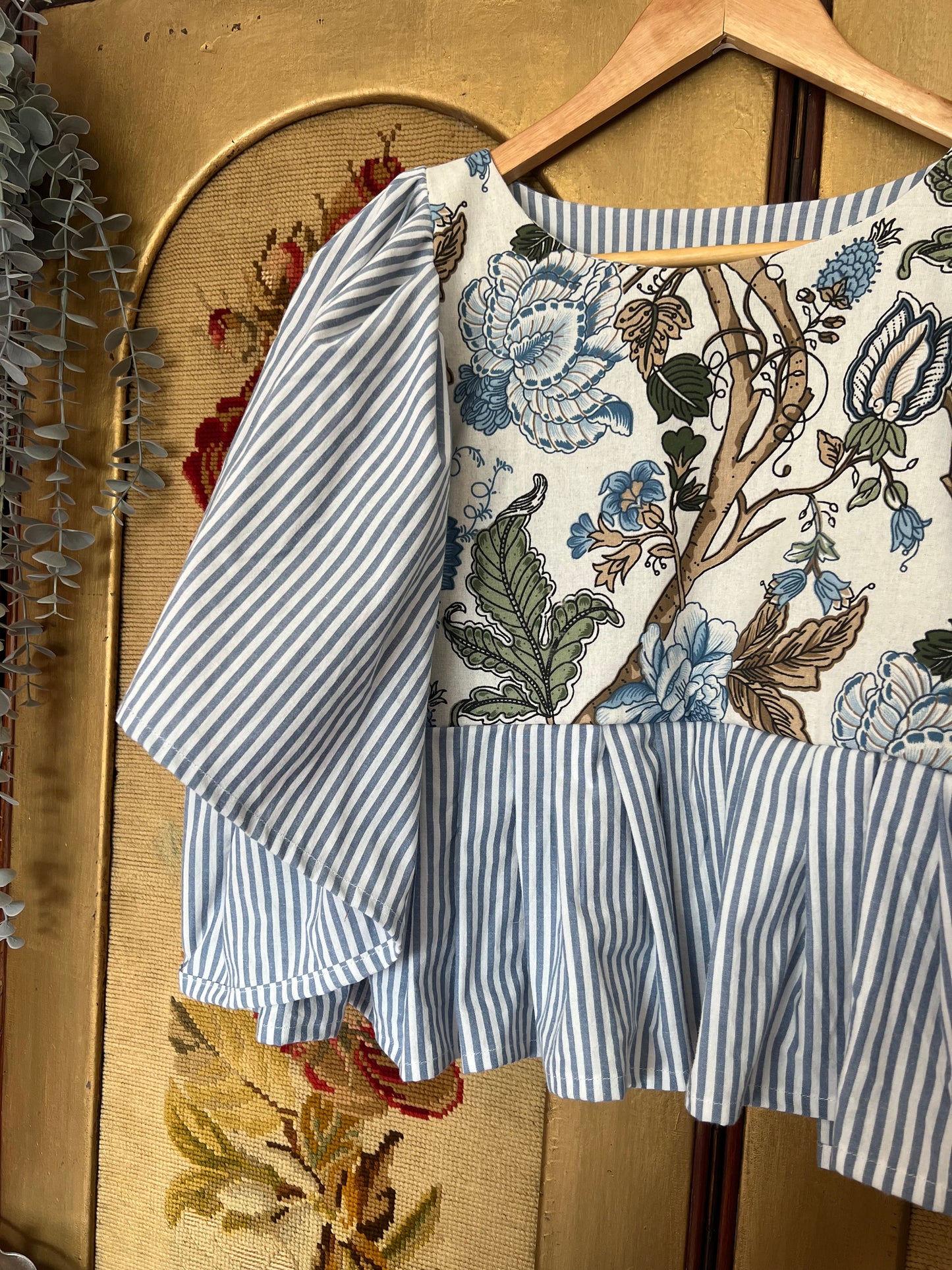 Floral-patterned top with blue and white striped sleeves on a hanger against a textured wall.