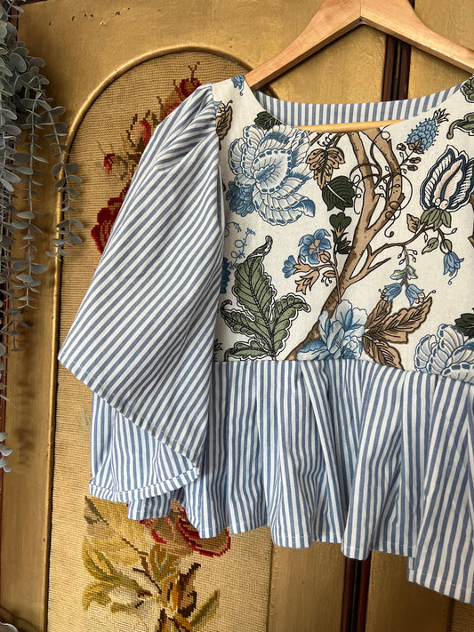 Floral-patterned top with blue and white striped sleeves on a hanger against a textured wall.