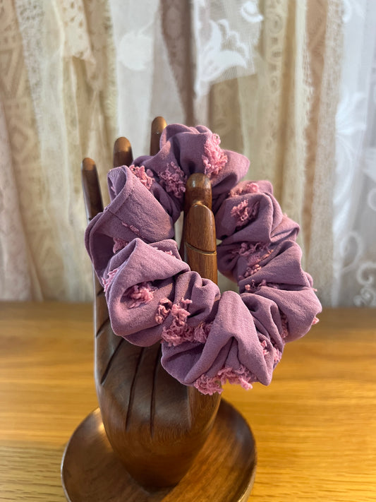 Purple scrunchies with floral details on a wooden holder against a lace curtain background.